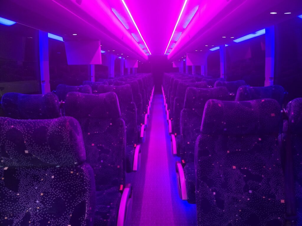 Interior of a bus illuminated with vibrant purple and blue lights, showcasing plush, patterned seating rows creating a modern, ambient atmosphere.