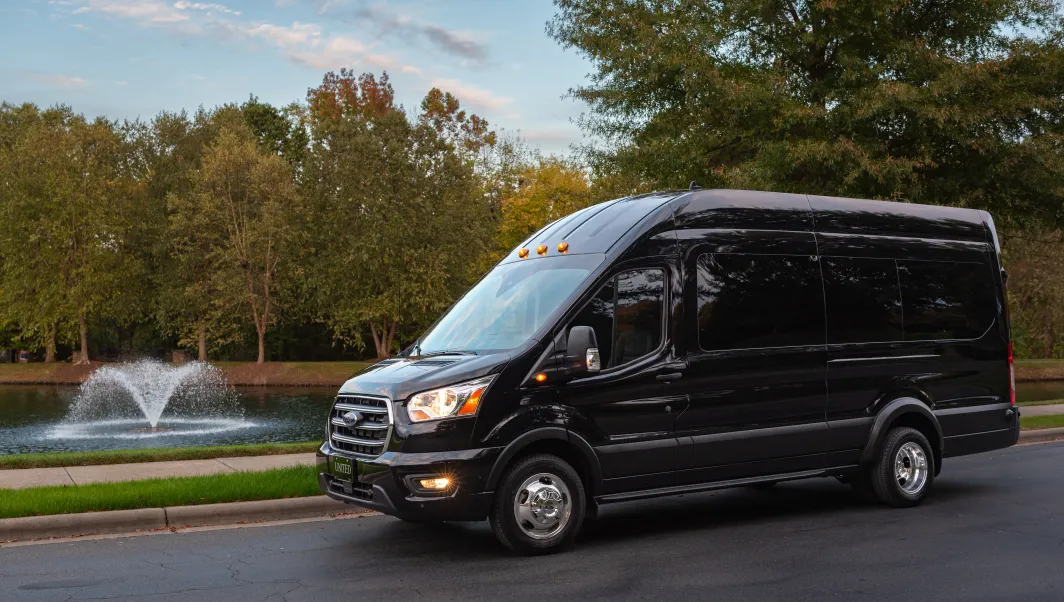 A black shuttle van parked near a pond with a fountain, surrounded by trees, showcasing its sleek design and spacious build.