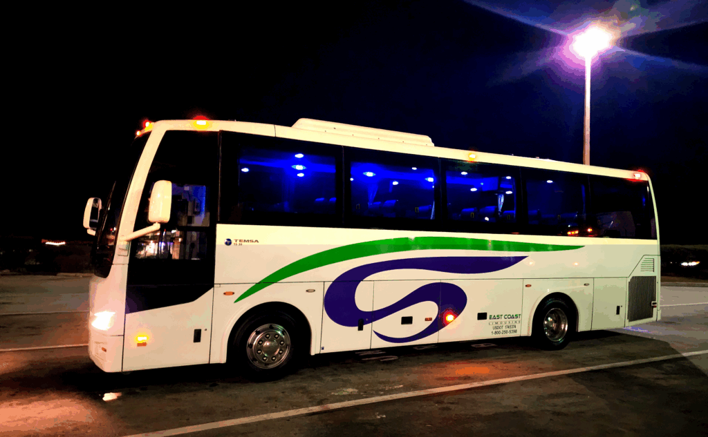 A modern white bus with green and purple accents is parked at night, illuminated by blue interior lights and street lamps, ready for passengers.