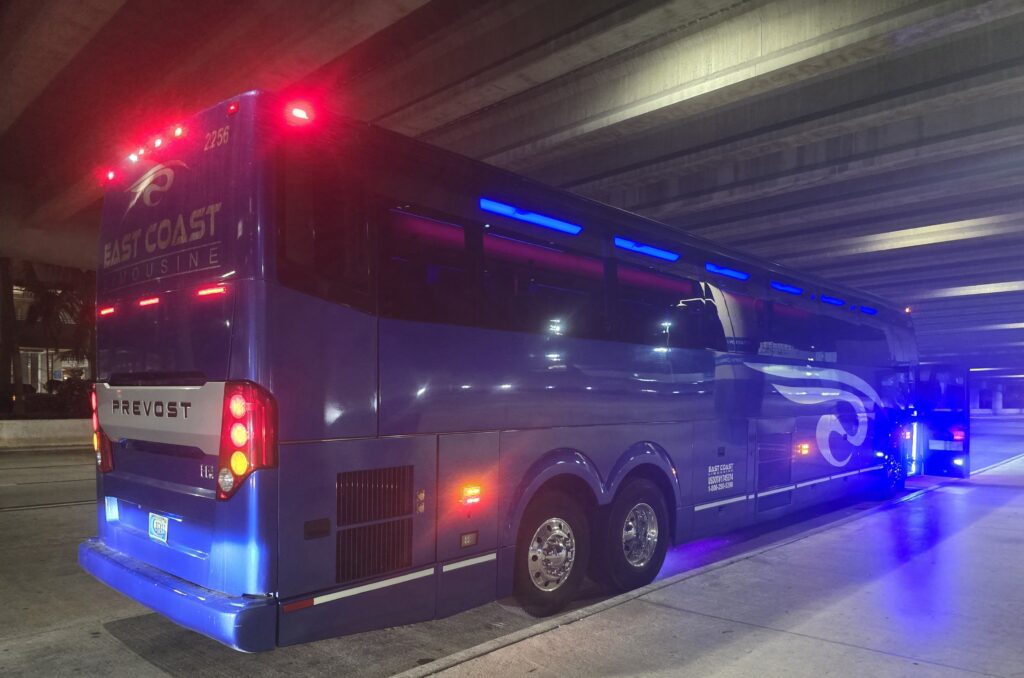 Blue East Coast Limousine bus parked under a highway overpass, illuminated with blue lights. The bus features the Prevost logo and is ready for passenger boarding.