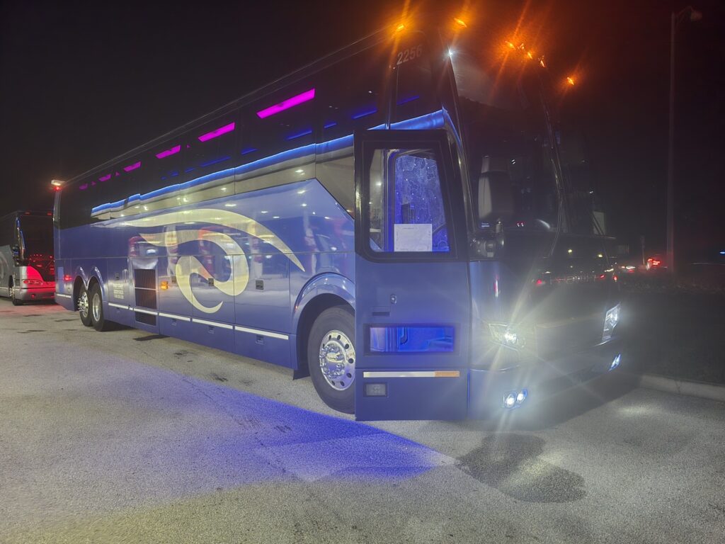 Blue luxury tour bus parked at night, illuminated by purple and white lights, showcasing sleek design and modern features.
