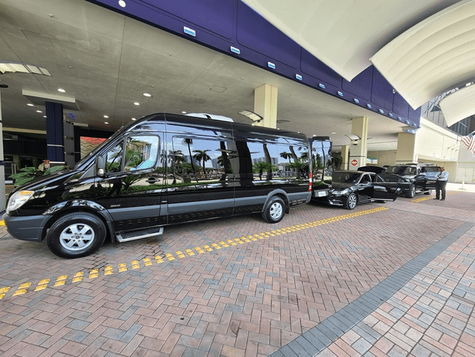 A sleek black shuttle bus and an accompanying luxury sedan wait curbside outside a modern building with palm trees, indicating transport services.