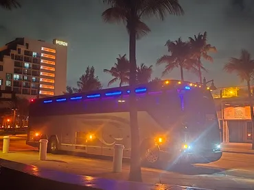 A sleek, illuminated bus parked along a palm-lined street at night, showcasing vibrant blue lights. Nearby buildings provide an urban backdrop.