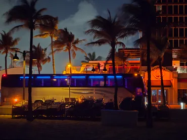 A brightly lit bus parked near a beach at night, surrounded by palm trees and lounge chairs, creating a vibrant coastal nightlife scene.