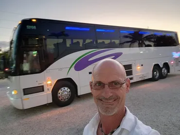 A man smiles for a selfie in front of a large, white tour bus with purple accents. The scene is set during dusk, suggesting travel or a tour event.