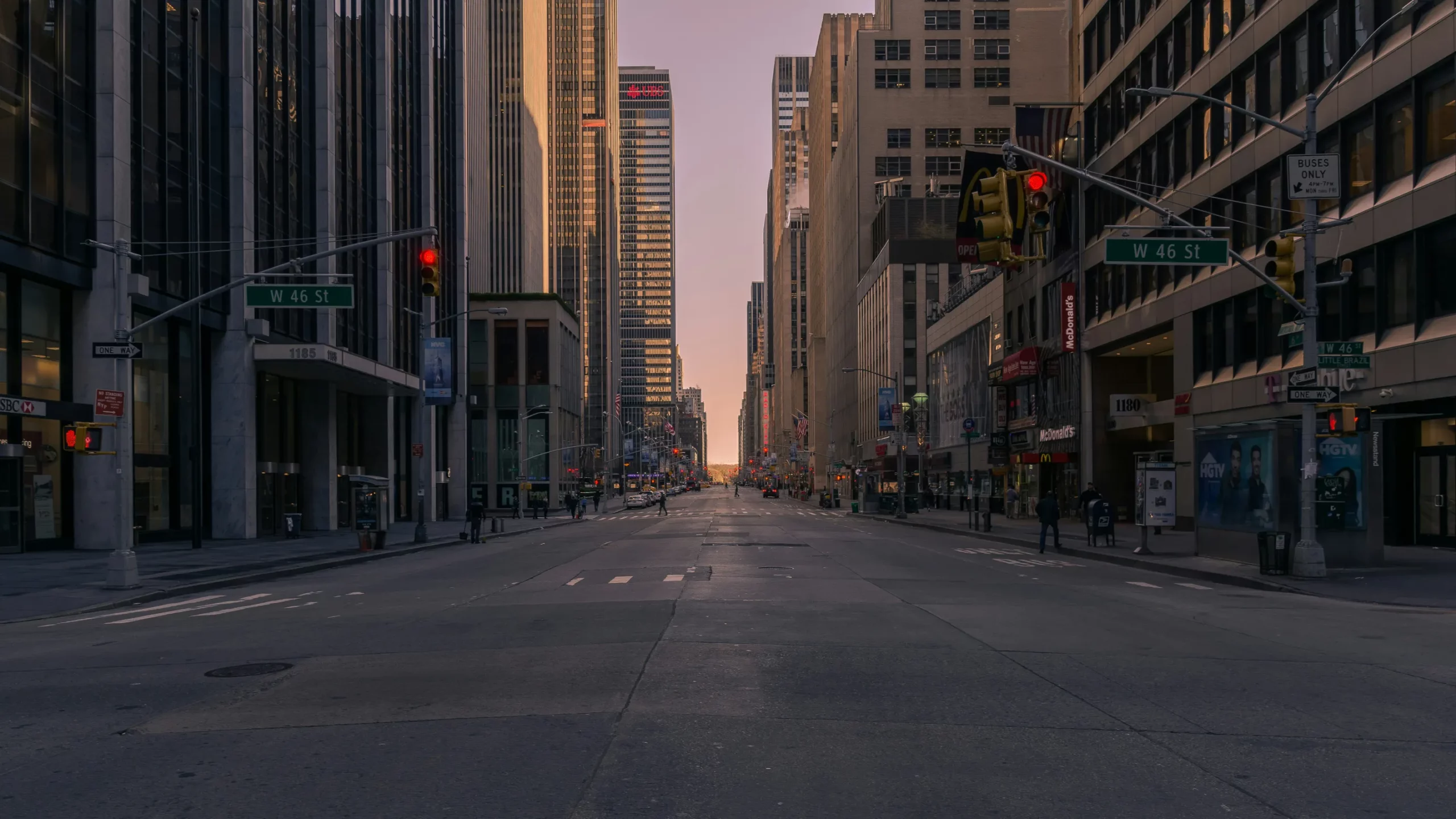 Quiet street view of W 46th St in New York City, flanked by tall buildings. Traffic lights display red; few pedestrians visible, emphasizing calmness.