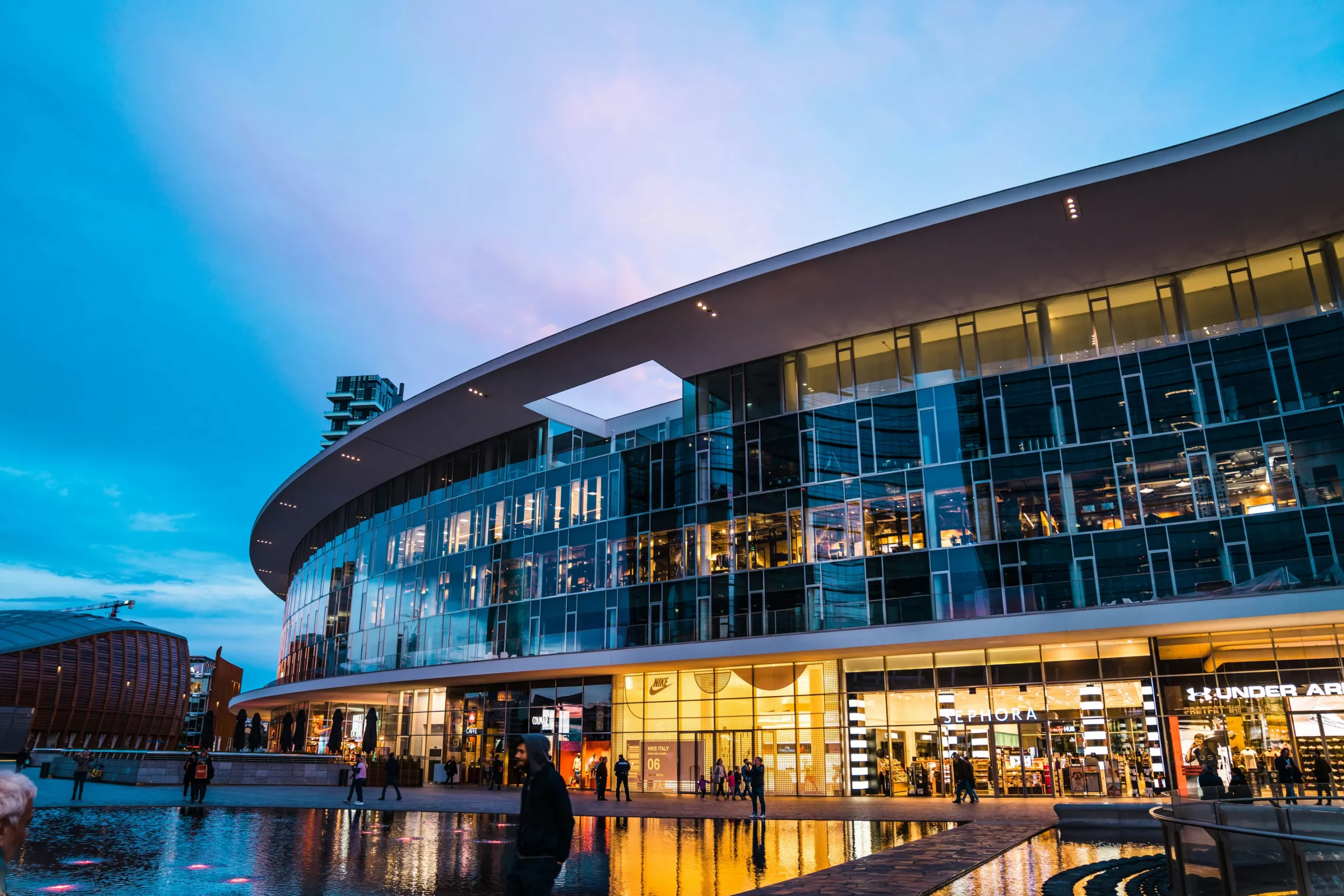 Modern shopping center featuring large glass windows and vibrant storefronts, reflecting dusk sky; people stroll nearby a tranquil water feature.