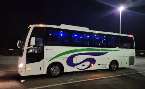 A white coach bus with a green and purple design, parked at night under bright lights, indicating it may be awaiting passengers.